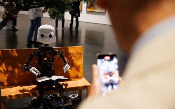 Zu sehen ist ein Roboter, dessen Aufbau dem Korpus des Menschen gleicht. Er sitzt auf einer Bank.
