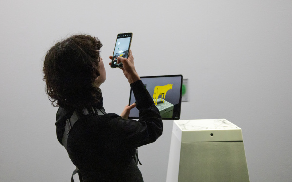 Ausstellungsansicht »Matter. Non-Matter. Anti-Matter« im ZKM | Zentrum für Kunst und Medien Karlsruhe, 2022. Eine Person fotografiert mit ihrem Handy ein Tablet, auf dem eine gelbe Kuh zu sehen ist.