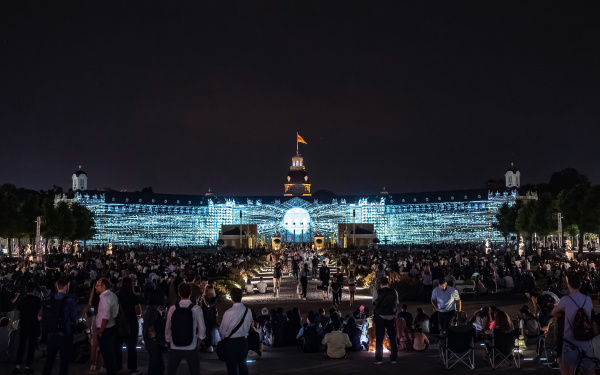 Das hell erleuchtete Schloss hebt sich stark von dem schwarzen Nachthimmel ab. Zu sehen ist etwas, das an eine Sonne erinnert, deren Strahlen sich über das Schloss erstrecken.