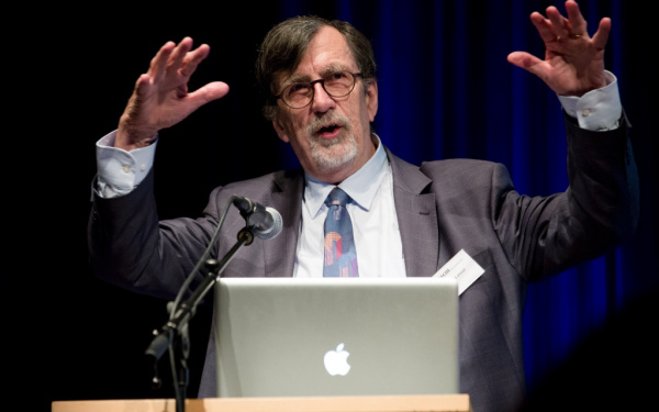 Bruno Latour vor einem Laptop und Mikrofon. Er hat beide Arme als Gestikulation in der Luft.