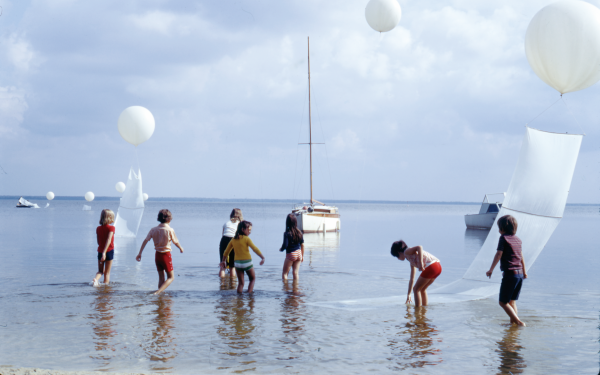 Zu sehen sind mehrer Kinder, die durch Wasser laufen. Über ihnen sind weiße Luftballons zu sehen. Im Hintergrund sieht man Boote. 