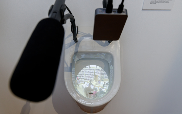 Zu sehen ist eine Toilettenschüssel, die von oben fotografiert wurde. In ihr befindet sich ein Projektion. Auf der Toilette ist ein Mikrofon befestigt.