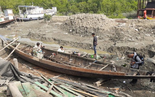 Man sieht ein Fischerboot, dass sich an Land befindet. Es sind vier Personen zu sehen, zwei davon arbeiten an dem Boot.