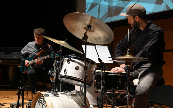 Zu sehen ist die Band »Polytheistic Ensemble«. Felix Schrack spielt Schlagzeug, dahinter befindet sich eine weitere Person mit einer Gitarre.