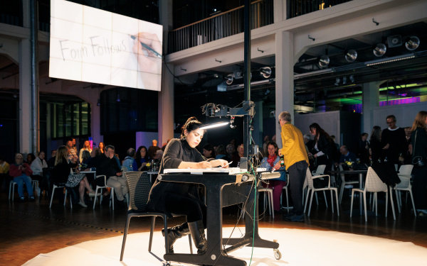 Im dunklen Foyer des ZKM ist in der Mitte von zuschauenden Menschen ein Tisch aufgebaut, der von einem Lichtkegel angestrahlt wird und an dem die Künstlerin Farzane Vaziritabar ihre Performance zeigt.