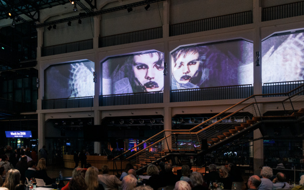 Das Foyer des ZKM | Karlsruhe ist voll mit Besuchern, die alle hoch an die Wand schauen, auf die ein Video projiziert wird. Auf dem Video zu sehen sind zwei Gesichter.