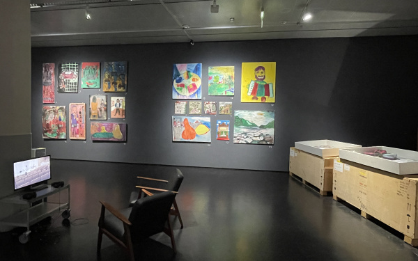 Ausstellungsansicht aus der Ausstellung "Wenn der Krieg ins Museum kommt". Zu ist ein Raum an dessen Wand viele der gemalten Bilder hängen und links ins eine Zone mit Schuhen und einem Computer, während rechts zwischen Holzkisten stehen
