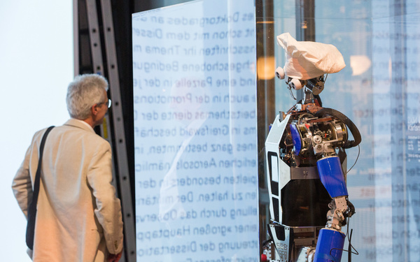 Zu sehen ist eine Person, die vor einem humanoiden Roboter steht. 