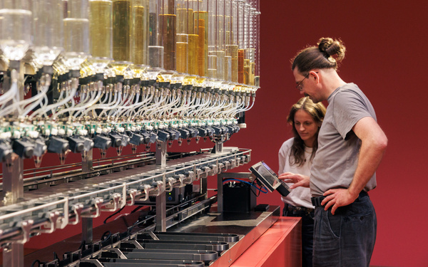 Two people standing in front of a machine with many perfume bottles.