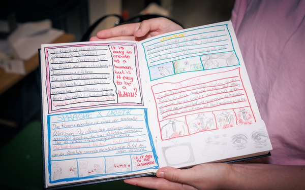 Das Foto zeigt ein aufgeschlagenes Notizbuch mit beschrifteten Seiten, das von einer Person in die Kamera gehalten wird.