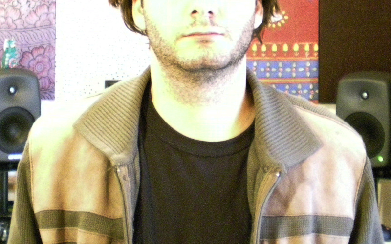 Ein Mann im T-Shirt und Jacke. Im Hintergrund zwei Lautsprecher vor einer farbig strukturierten Wand.