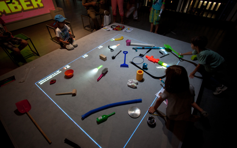 Kinder sitzen vor einer Spielfläche auf der Gegenstände liegen und virtuelle Autos fahren