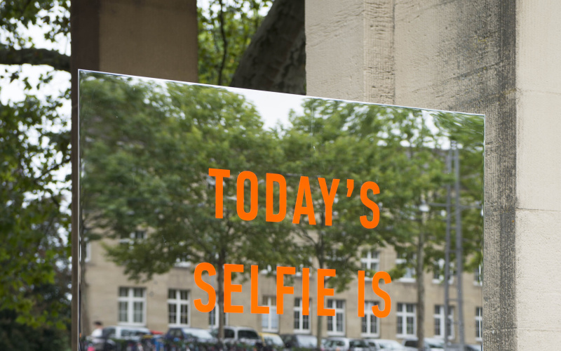 An orange lettering "Todays Selfie is Tomorrows Biometric Profile" can be seen on a large rectangular frameless mirror. The installation stands outside in front of the entrance south of the zoo.