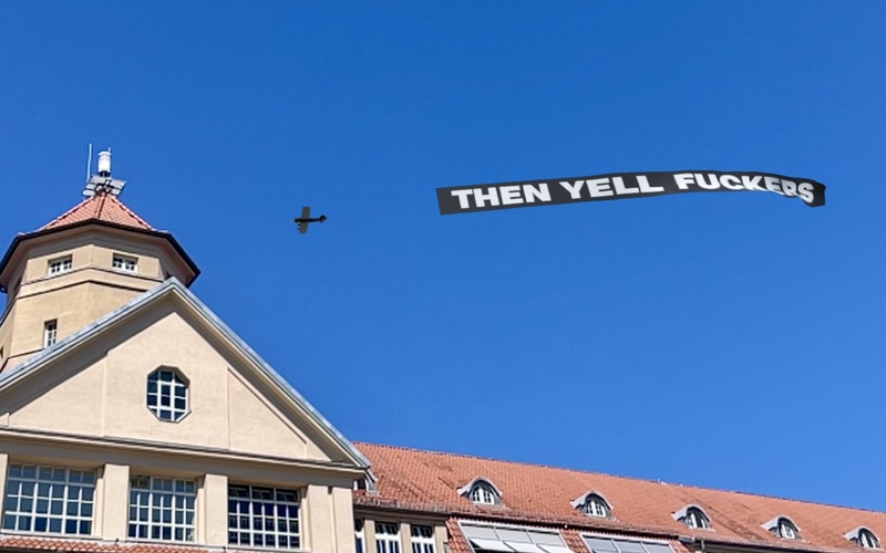 Zu sehen ist ein Flugzeug am Himmel über dem ZKM, das ein schwarzes Banner mit Schrift hinter sich herzieht.