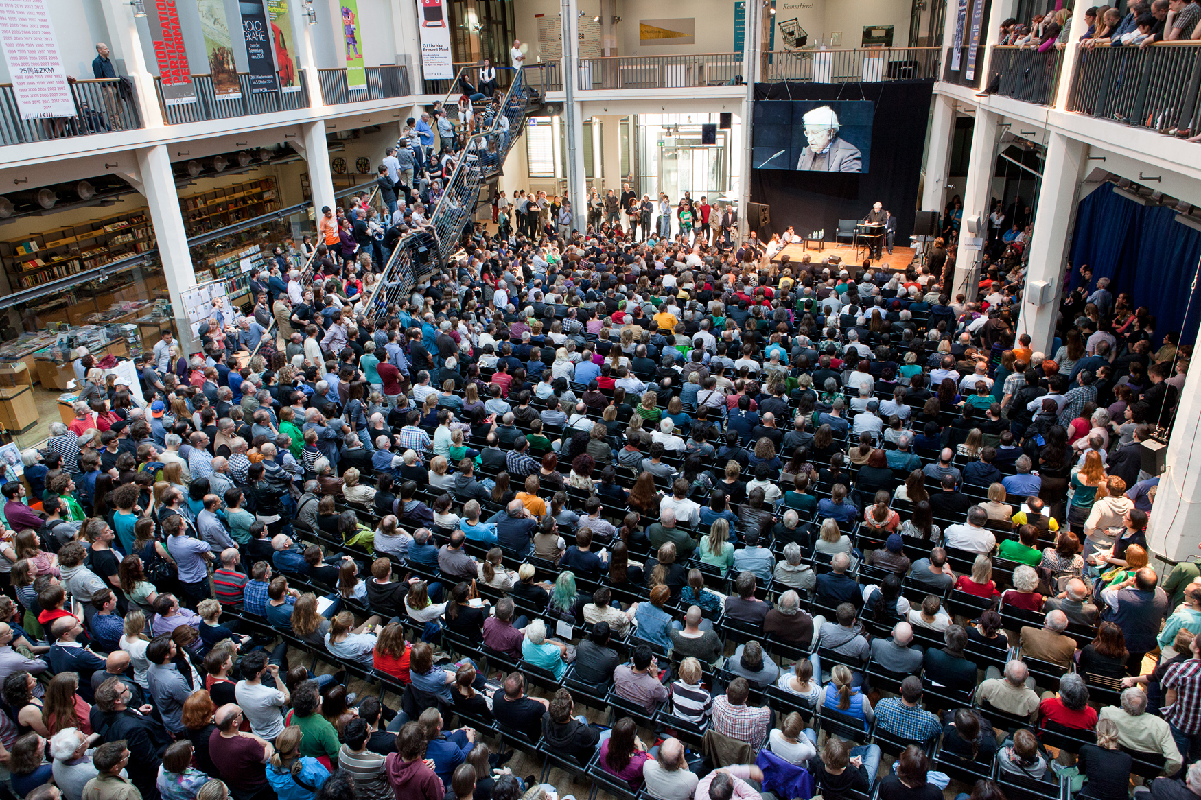 In front of a stage on which a man gives a lecture, many people sitting on chairs