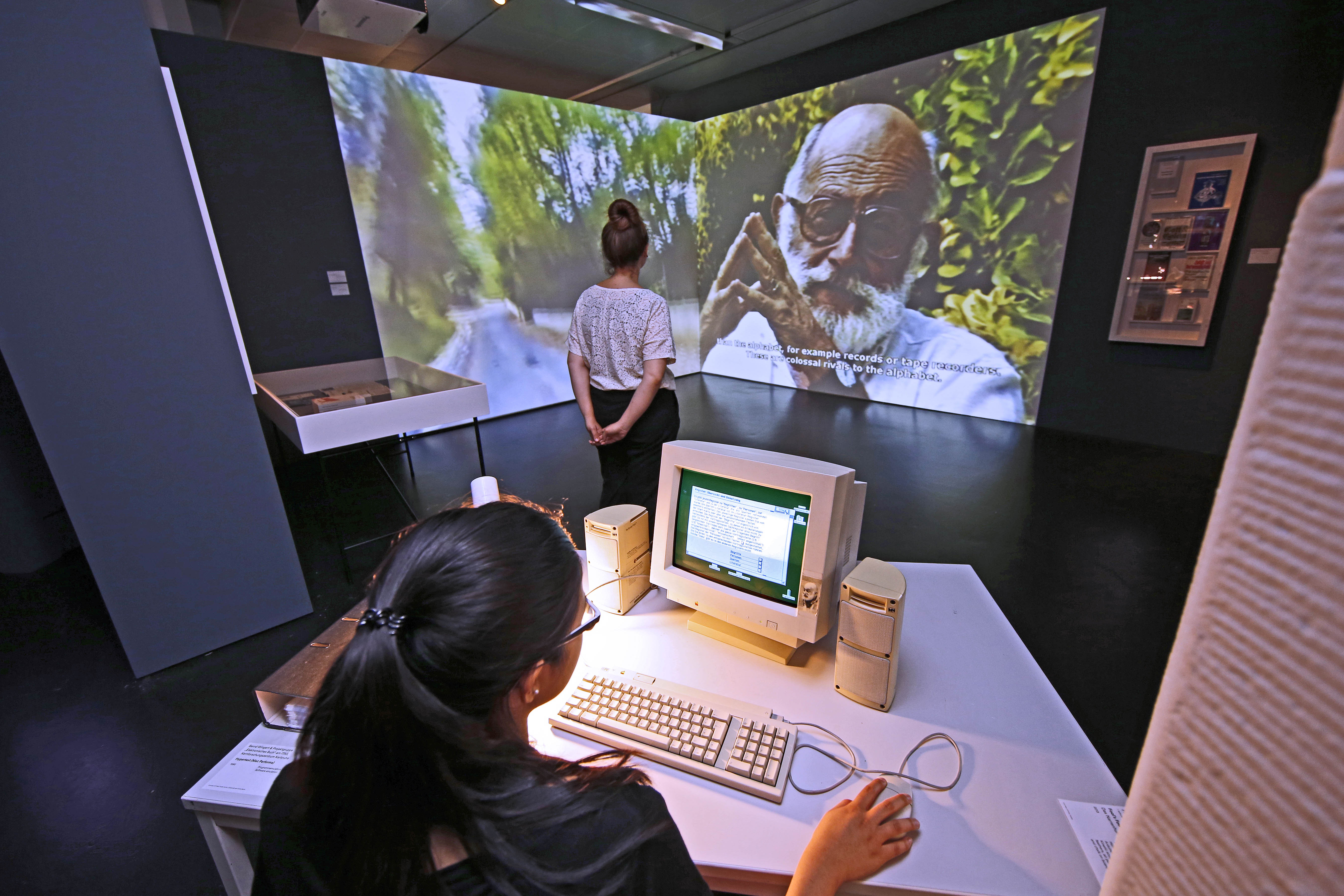 Exhibition view: foreground a girl sitting at a computer, in the background a video projection with Vilém Flusser that fills an entire wall