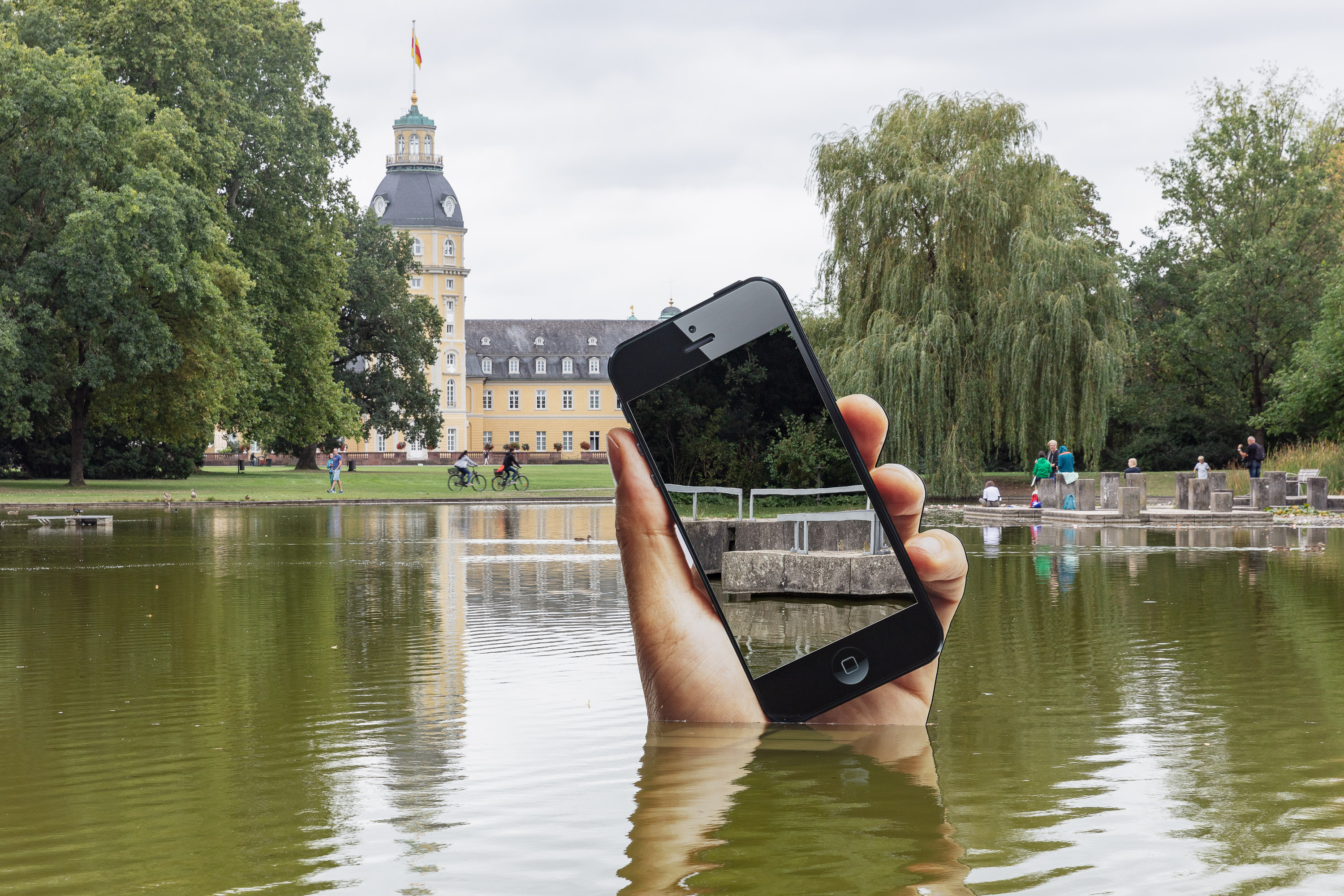 Zu sehen ist das Ufer eines Sees. Aus dem See heraus ragt eine sehr große Hand mit einem Smartphone. Das Display des Smartphones ist ein Spiegel, in dem sich die Betrachter am Ufer sehen können.