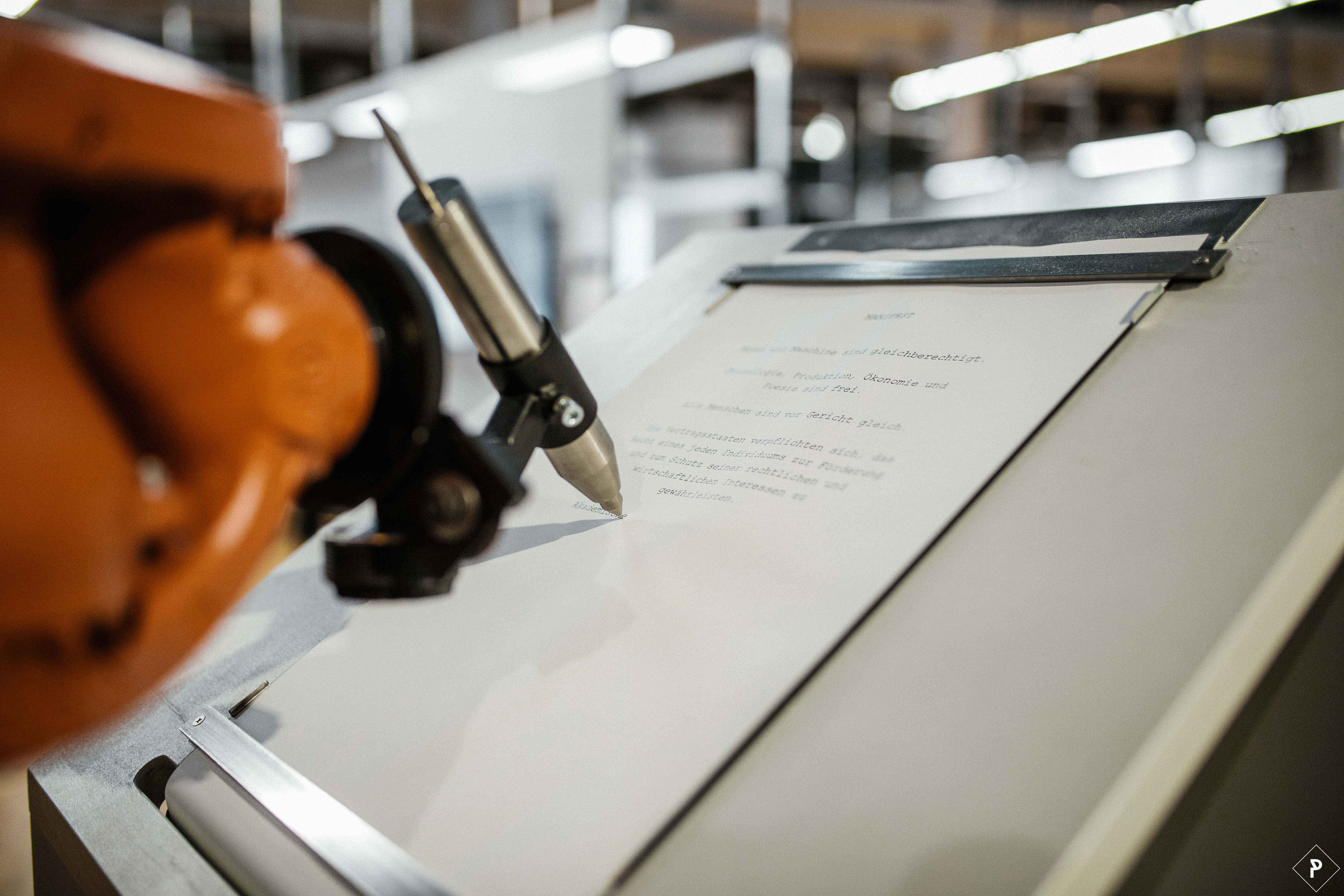 Ein orangefarbener Roboterarm schreibt mit einem silbernen Stift auf einem Blatt Papier