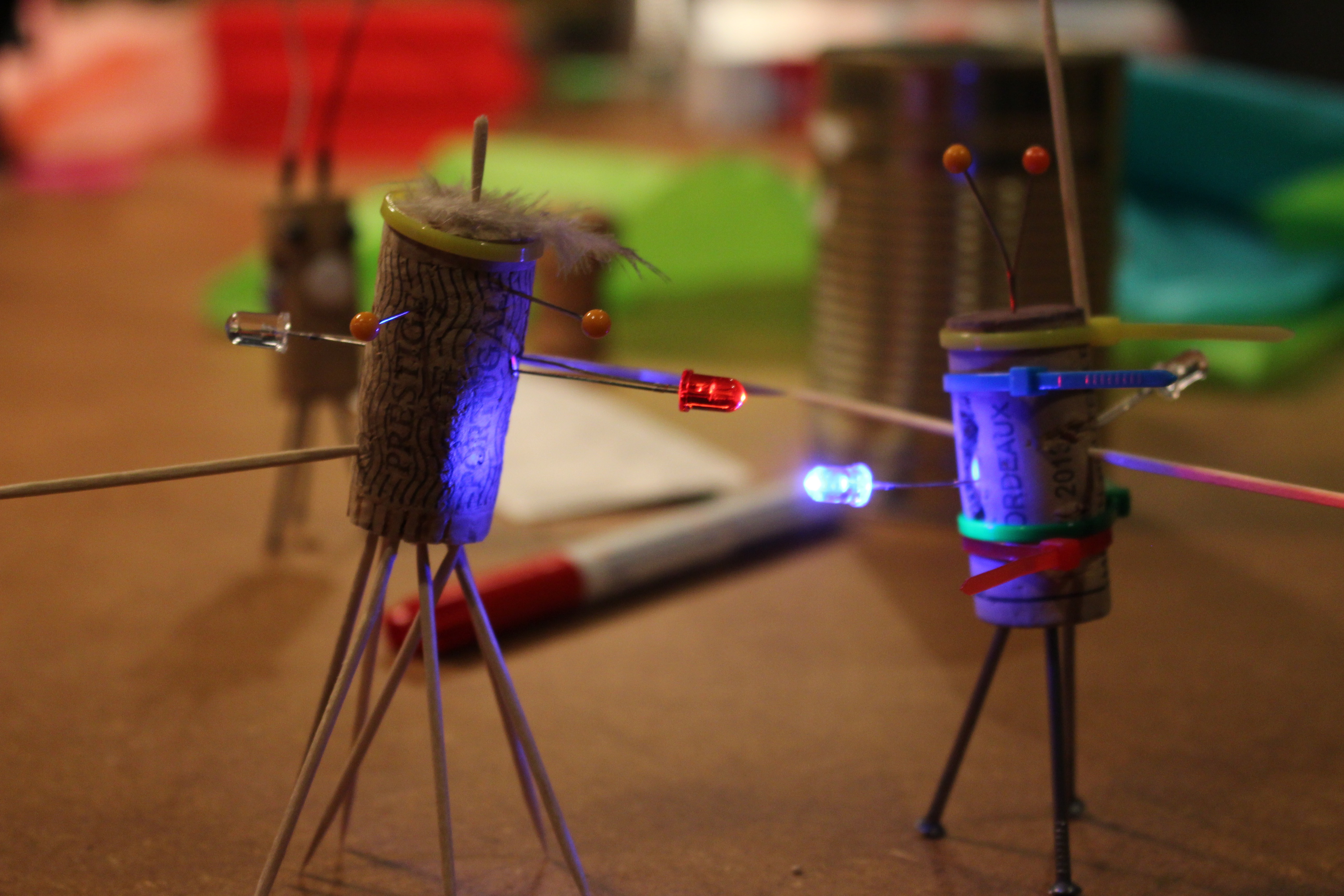 Zwei Korken, die zu kleinen Figuren umgewandelt wurden und mit LEDs ausgestattet sind stehen auf einem Tisch mit allerlei Bastelutensilien.