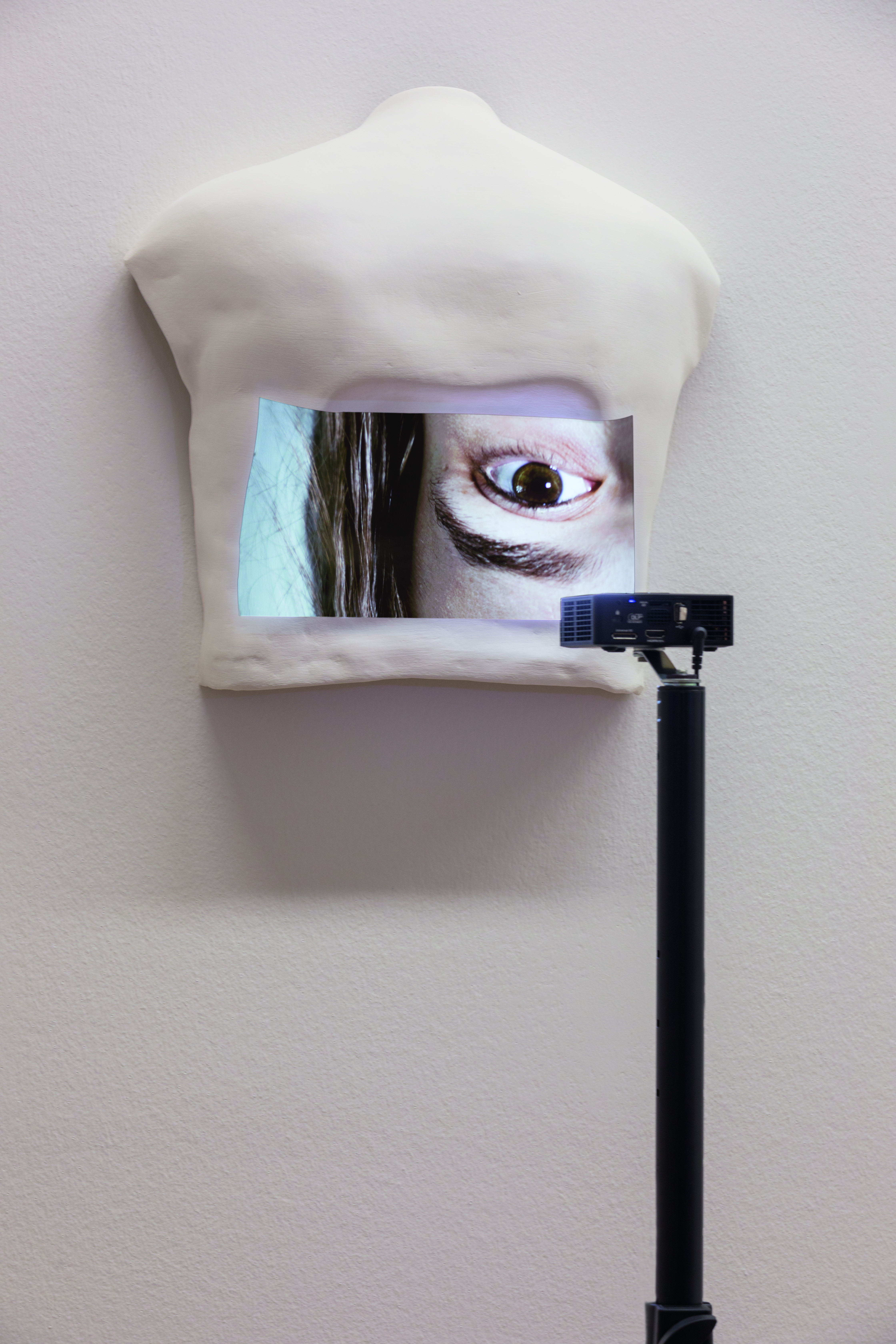 Zu sehen ist ein weißes Objekt, welches an der Wand hängt. Auf dem Objekt ist das Bild eines Abschnitts aus dem Gesicht einer Frau. 