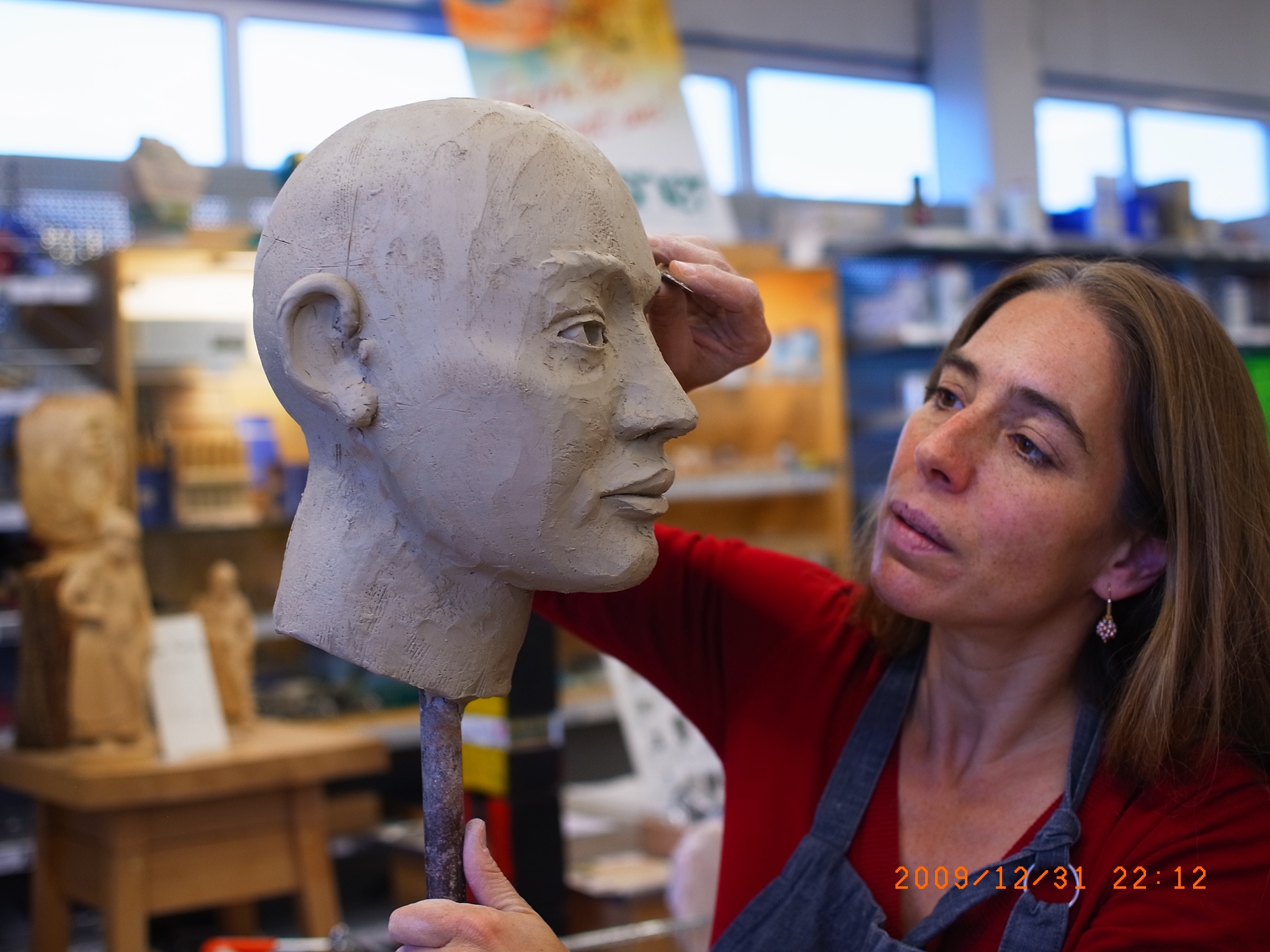 A model of a head sticking on a pole. A woman is working on it with a sculpting tool.