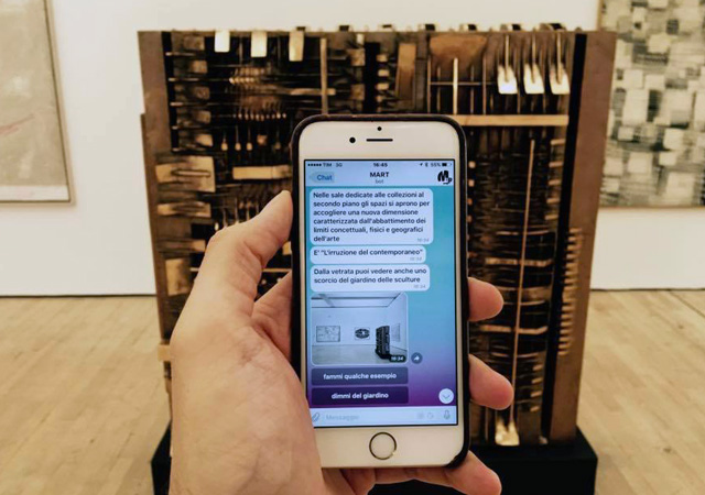 Smartphone in the hand of a museum visitor, with chat messages from the Bot Mart, in front of an artwork in the museum