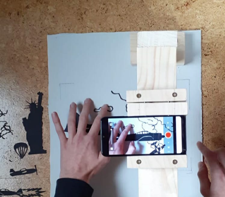 Das Bild zeigt als Nahaufnahme eine Hand, verschiedene Materialien und Schablonen sowie ein Mobilgerät. Das Bild ist im Rahmen der Kulturakademie Baden-Württemberg 2020/21 entstanden.