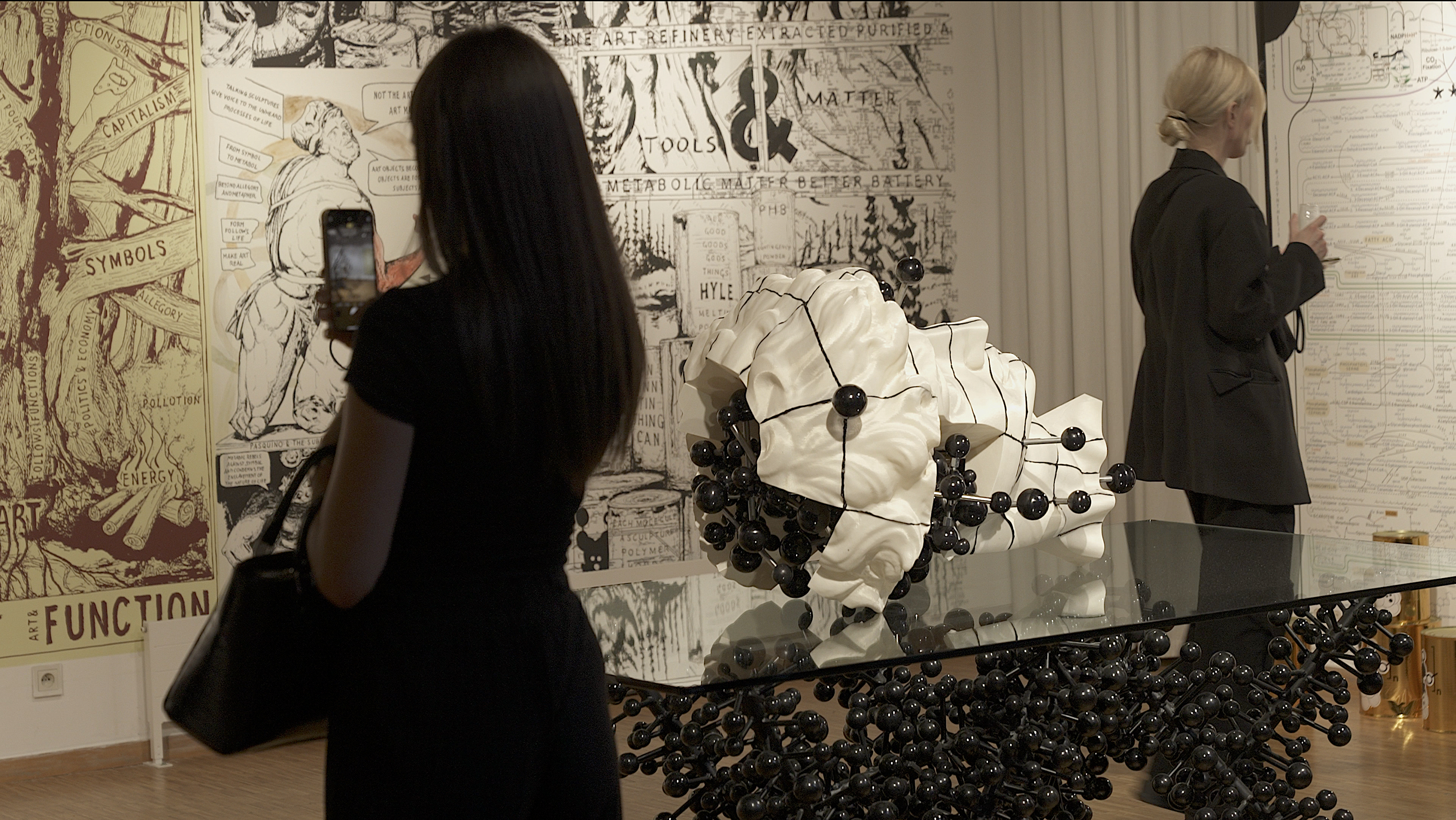 Two women walk through the exhibition. In the centre is a white sculpture of a head studded with black spheres
