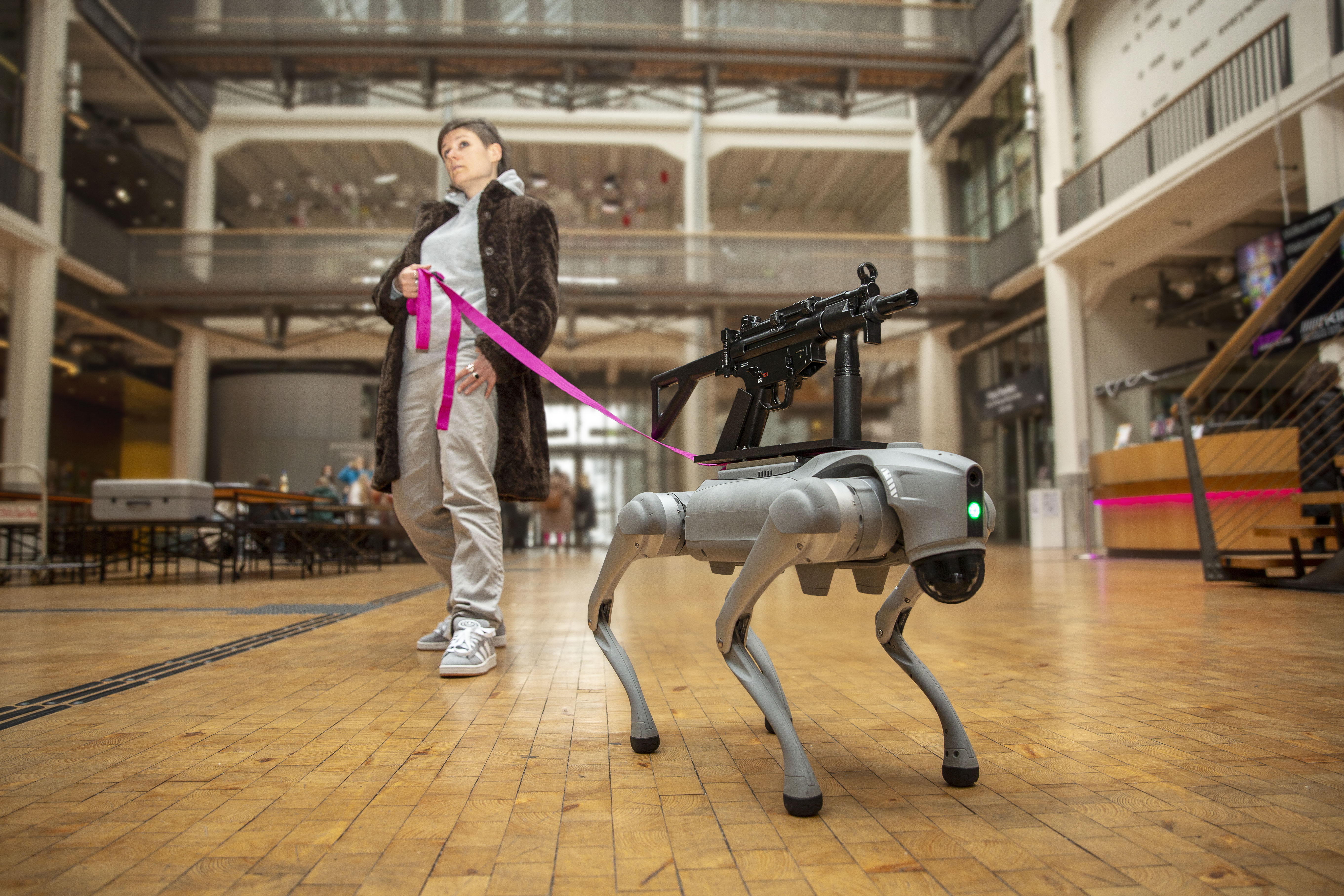 Ein Roboter Hund mit einem Sturmgewähr auf dem Rücken, der von einer pinken Leine von einer Person mit braunen Haaren und Pelzmantel gehalten wird.