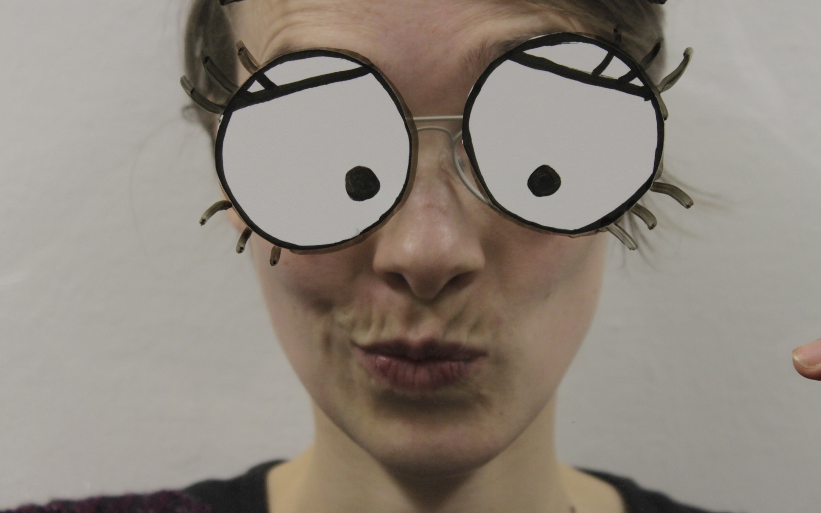 Eine junge Frau schaut verschmitzt mit gekräuselten Lippen in die Kamera. Ihre großen comic-artigen Augen sind auf eine Folie aufgezeichnet, die sie vor ihr Gesicht hält.