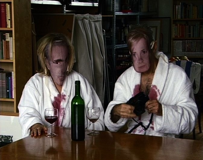 Zwei Personen mit Masken trinken Rotwein. Ihre Bademäntel sind dabei voller Rotweinflecken.