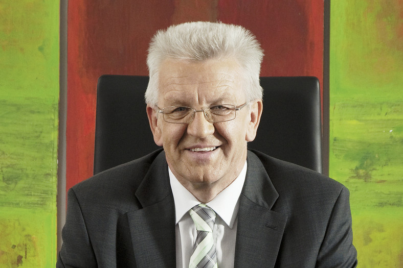 A laughing elderly gentleman. He is wearing a gray suit and a white shirt and a pair of glasses. His hair is gray. He sits on a black office chair. In the background, the colors are red and green to be seen.