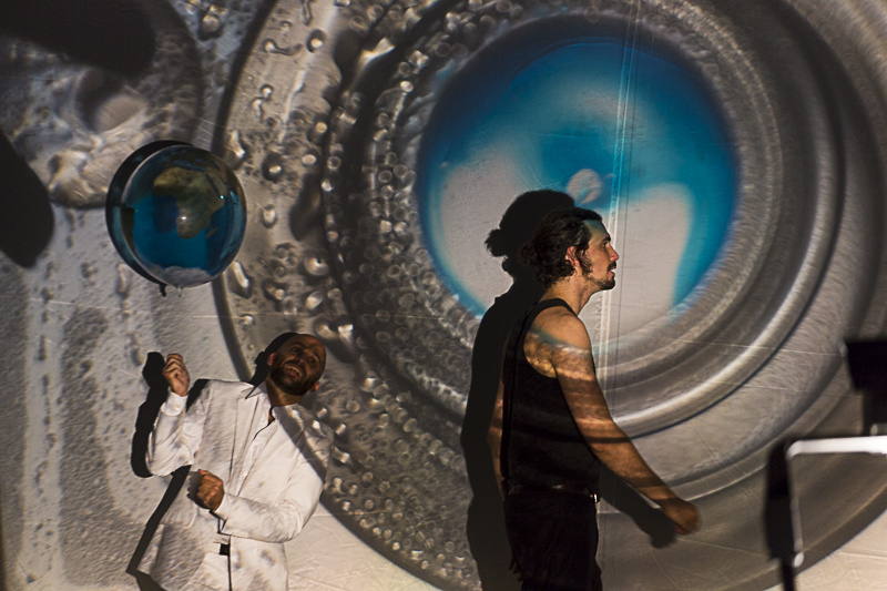 A man dressed in black standing in profile in front of a large silver circle element whose center is bluish. Behind him a man dressed in white holding a balloon in his hands.