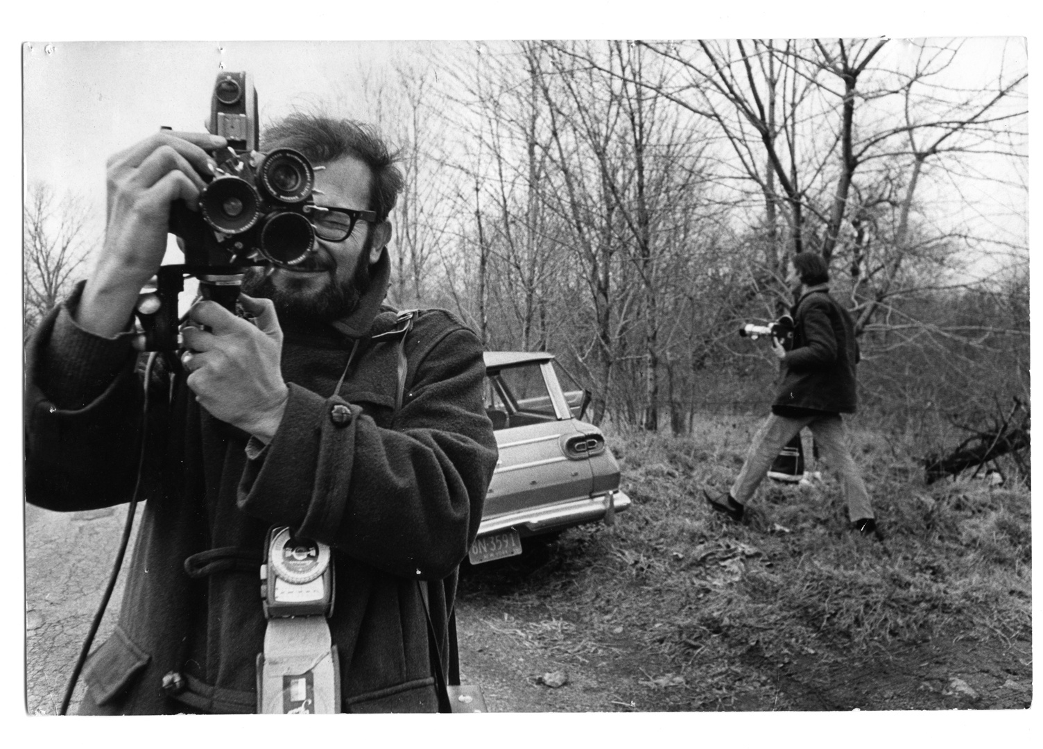Ein schwarz-weiß Bild von einem Mann, der ebenfalls fotografiert. Im Hintergrund steht ein Auto und ein weiterer Mann läuft in das Bild