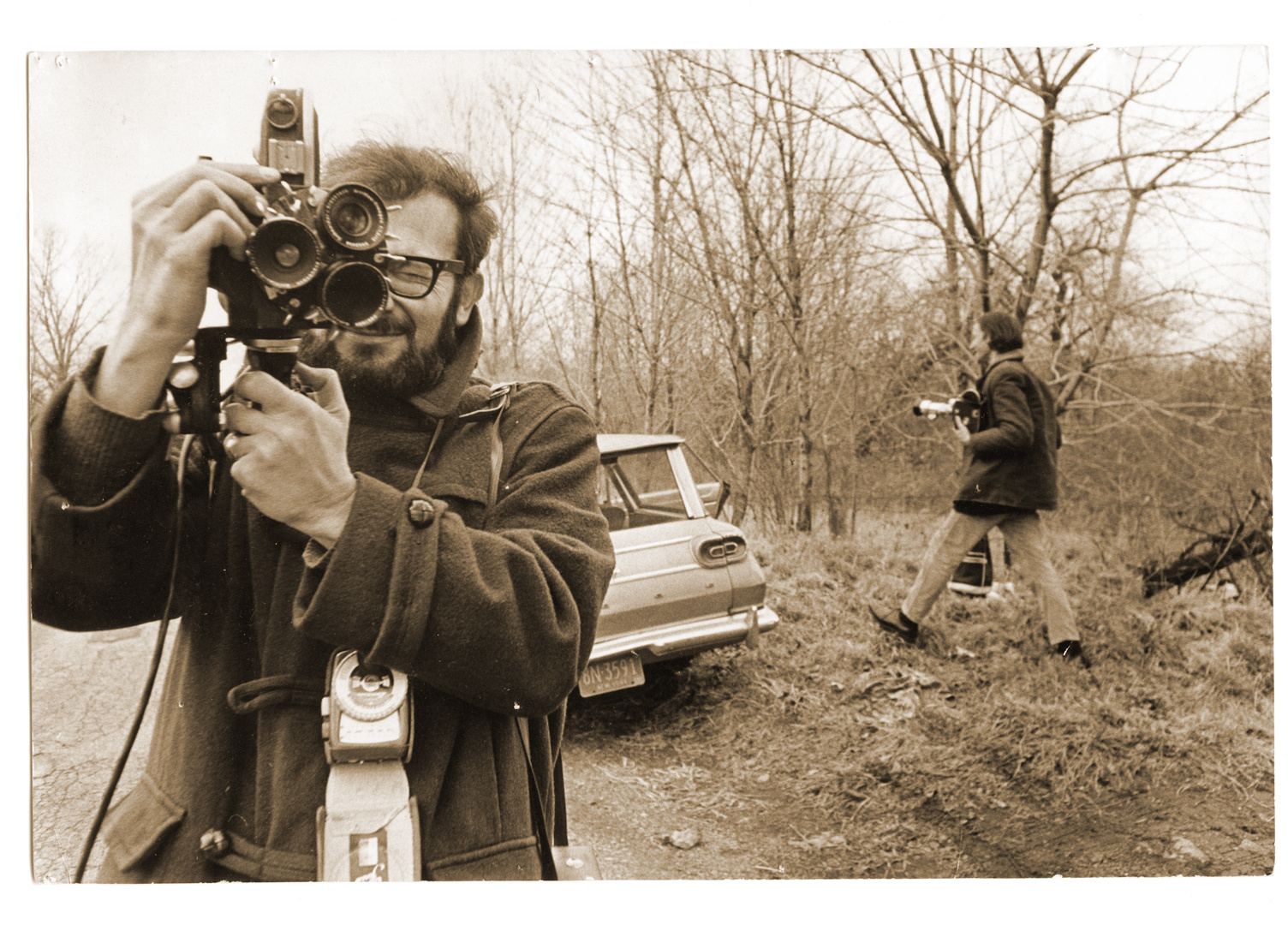 Ein schwarz-weiß Bild von einem Mann, der ebenfalls fotografiert. Im Hintergrund steht ein Auto und ein weiterer Mann läuft in das Bild