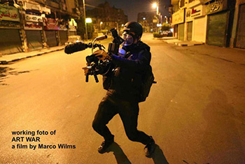 A road at night. In the foreground a man dressed in black with a helmet and protective mask. Equipment in his hands.