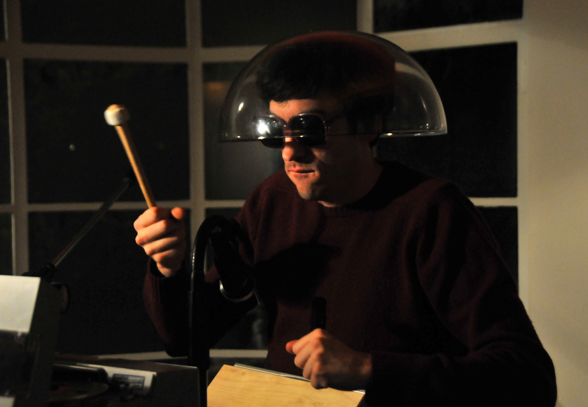 Ein Mann mit Sonnenbrille und durchsichtigem Lampenschirm auf dem Kopf, der einen Drumstick in seiner rechten Hand hält.