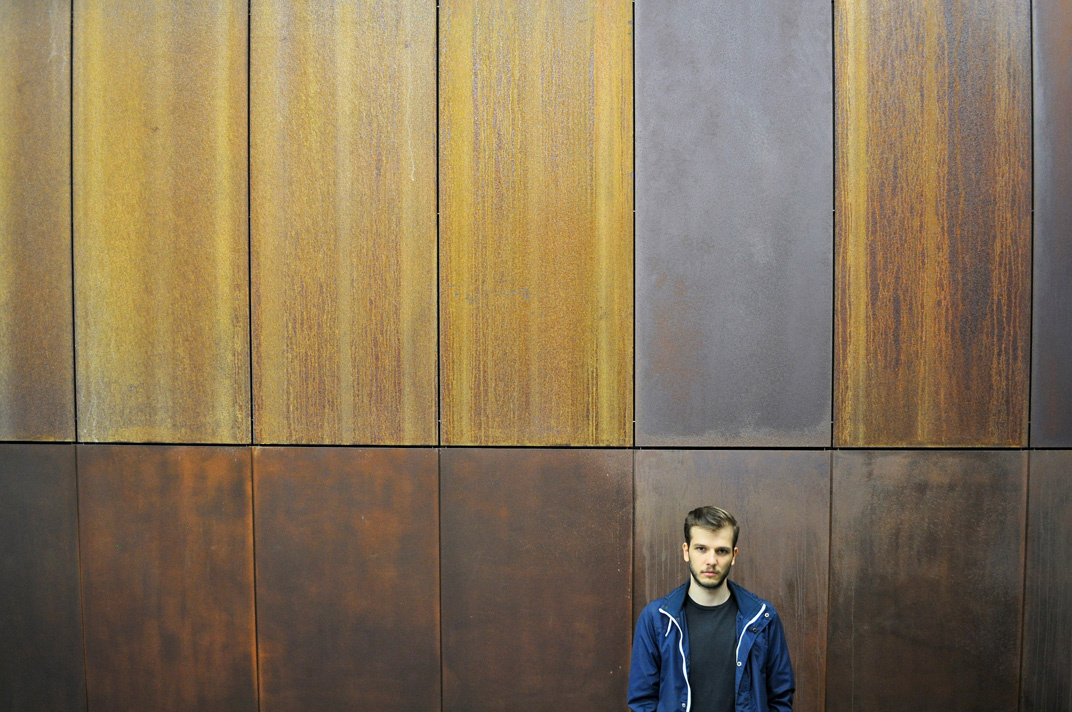 Ein junger Mann mit blauer Sportjacke steht vor einer Holzwand