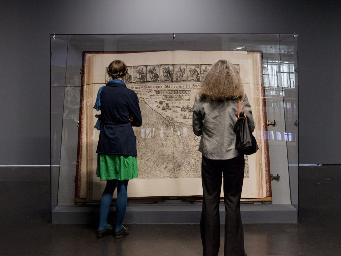 Der Klencke Atlas (1660) ist einer der größten Atlanten der Welt. Aufgeschlagen steht er in einer ihn schützenden Glaskonstruktion. Zwei Frauen in Rückenansicht betrachten ihn.