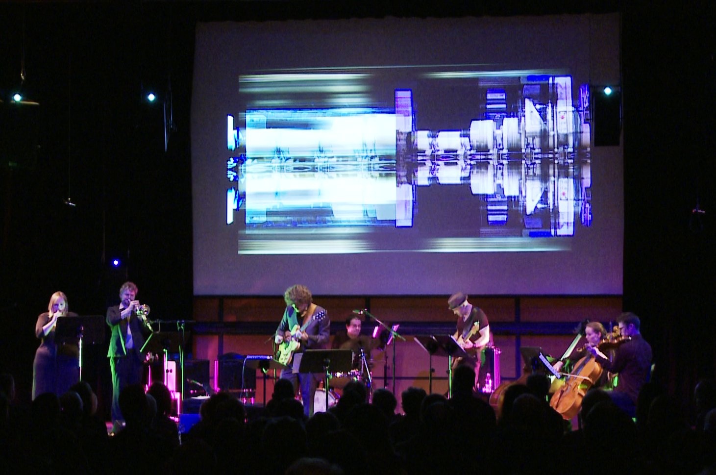 Musizierende Band auf einer Bühne. Über ihnen im Hintergrund futuristische Videoanimationen.