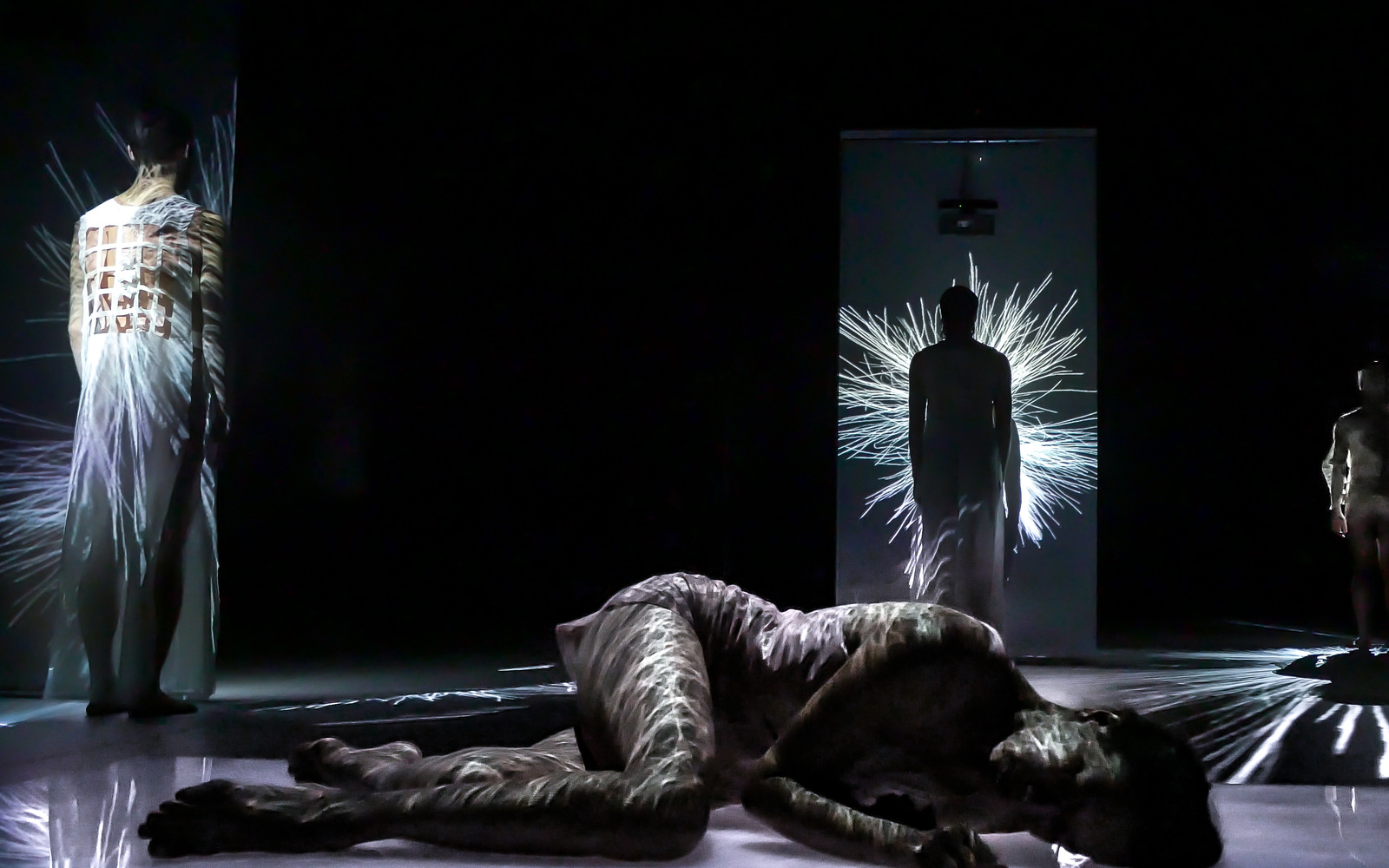 Four dancers are on stage, it is dark. One dancer is at the front of the stage, the others are standing at a distance to one direction each. On all dancers you can see numerous threads of light projected.