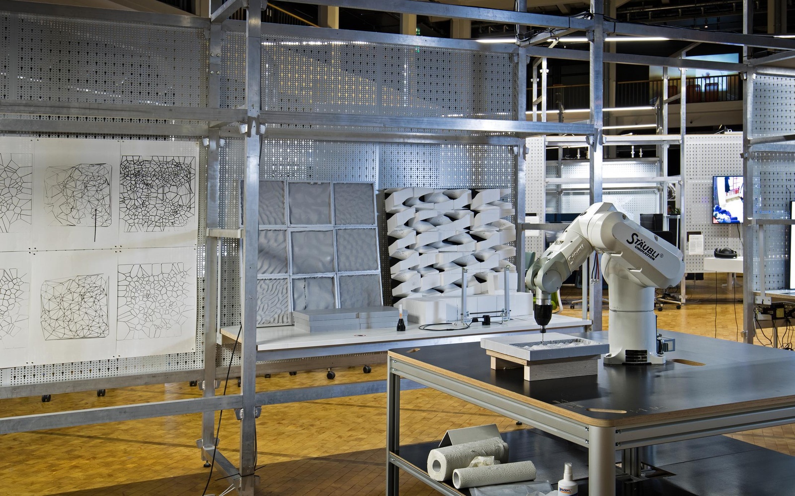 Im Vordergrund steht ein Roboterarm auf einem Holztisch, während man im Hintergrund verschiedene Plastiken sowie Zeichnungen an einer Gitterstruktur sieht