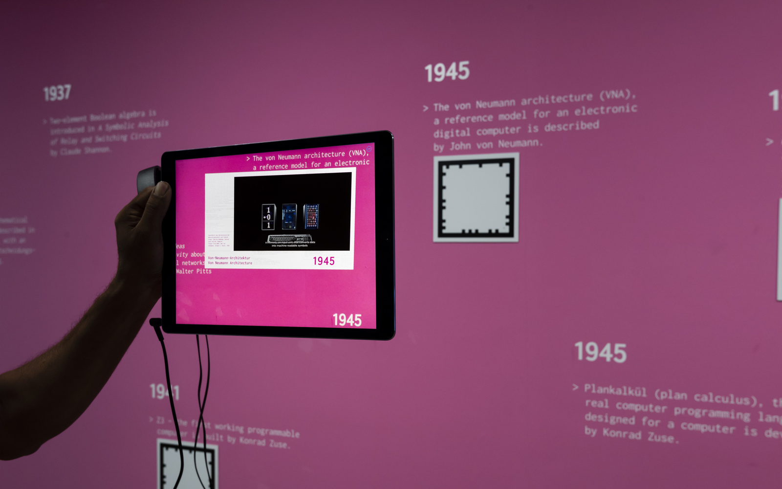 Eine Hand hält ein Tablet vor eine pinkfarbene Wand. 