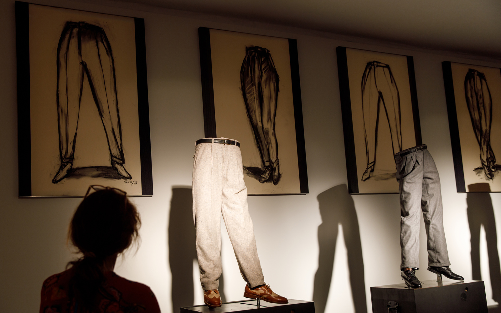 Foto einer Kunstinstallation: zu sehen sind Hosen mit Schuhen auf einer Art Laufsteg, im Hintergrund sind gerahmte Zeichnungen.