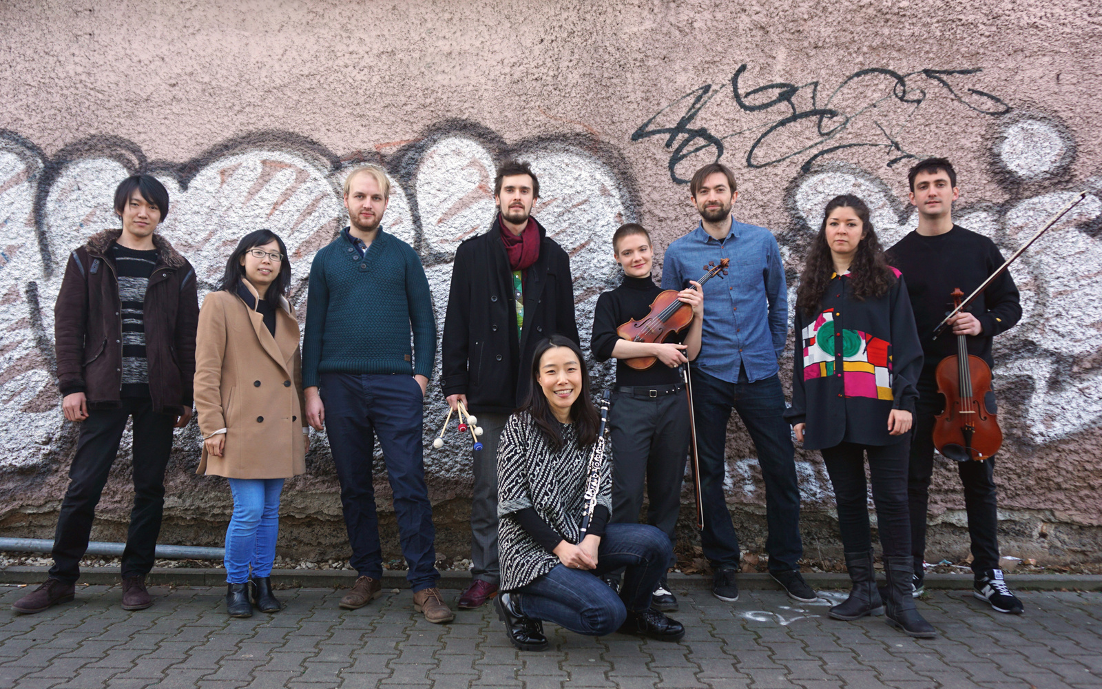 Das Bild zeigt die StipendiatInnen der Internationalen Ensemble Modern Akademie (IEMA) vor einer vollgesprühten Backsteinwand