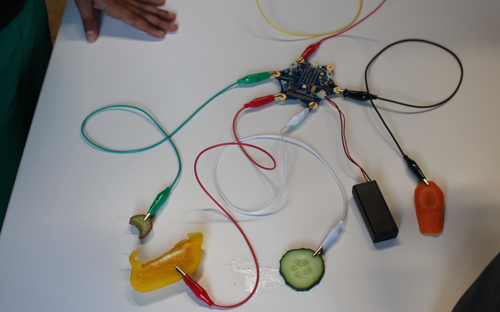 Wir sehen einen Calliope mini, von dem mehrere Kabel weg führen. An deren Enden wiederum verschiedenes Gemüse angeschlossen ist. 