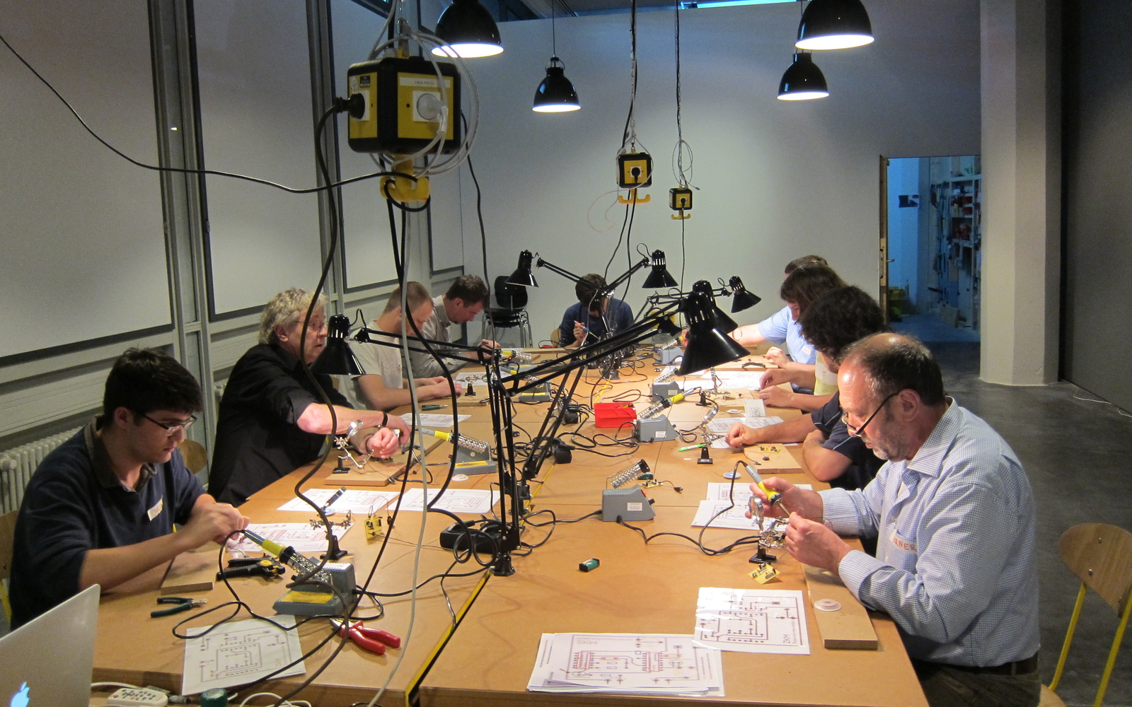 An einem riesigen Tisch sitzen viele Workshopteilnehmer, die mit Lötkolben arbeiten und es liegt allerlei Material kreuz und quer herum.