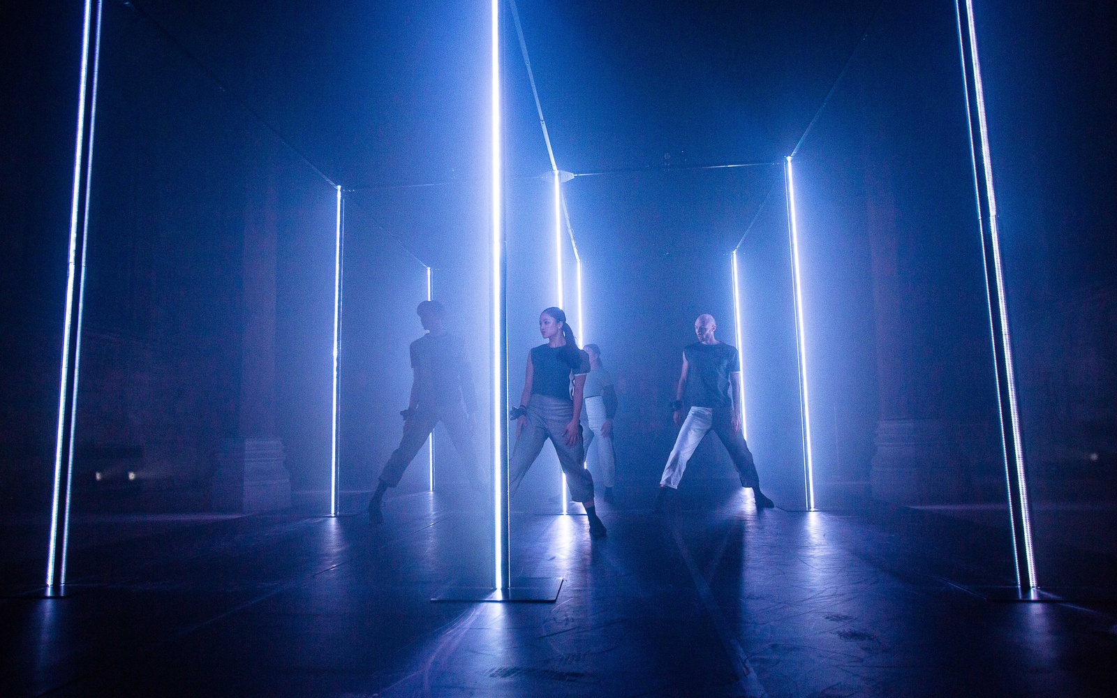 A group of dancers stands in a darkened room illuminated by blue neon tubes.