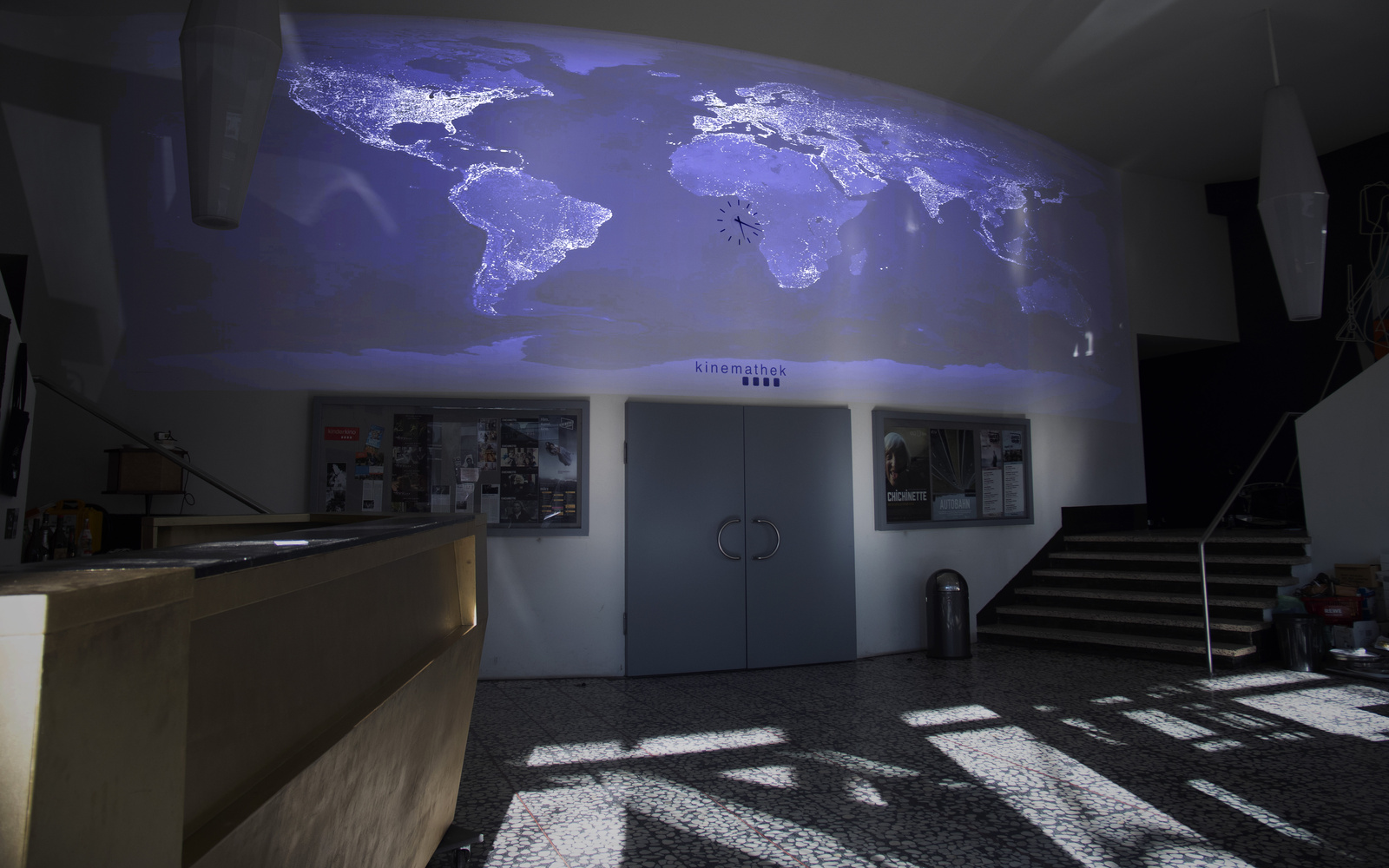 You can see a wall with a big door. Above the door is a world map projected onto the wall in cold colors.