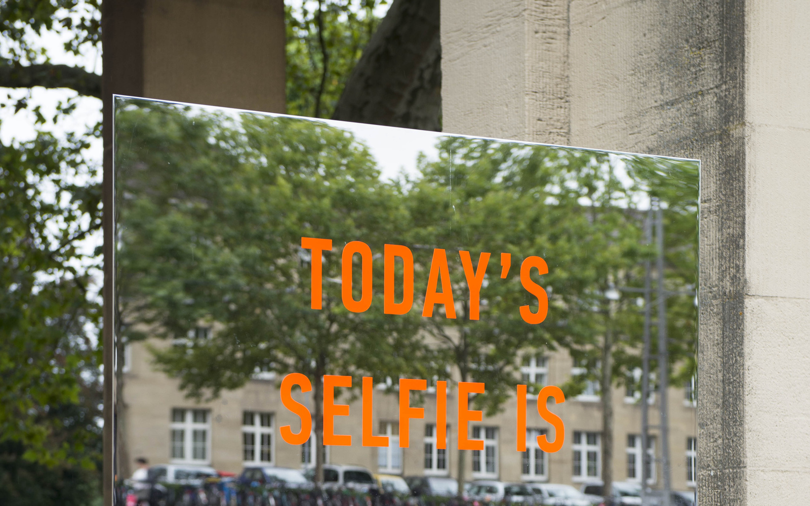 Zu sehen ist ein orangener Schriftzug »Todays Selfie is Tomorrows Biometric Profile« ist auf einem großen rechteckigen, rahmenlosen Spiegel. Die Installation steht draußen vor dem Eingang-Süd des Zoos.